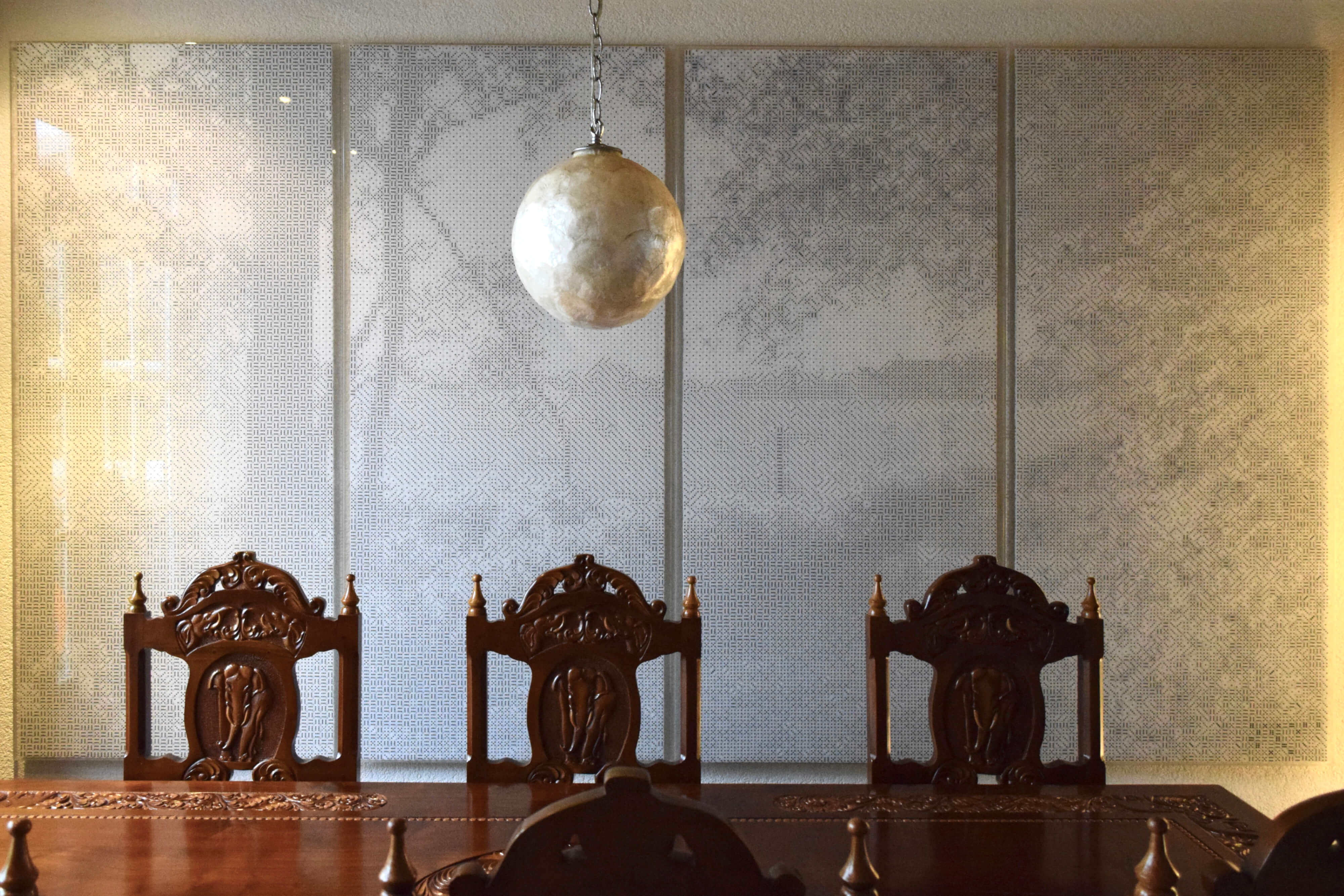 Een groot mozaïek van witte dobbelstenen met zwarte ogen boven een grote, klassieke, houten eettafel. Het mozaïek bestaat uit 4 langwerpige panelen, die samen een breed landschap vormen. Het onderwerp van het kunstwerk is een uitzicht over een weiland gezien vanaf een zandpad aan de bosrand. De lage zon schijnt tussen de bomen door en laat lange schaduwen van de begroeiing op het zandpad vallen.