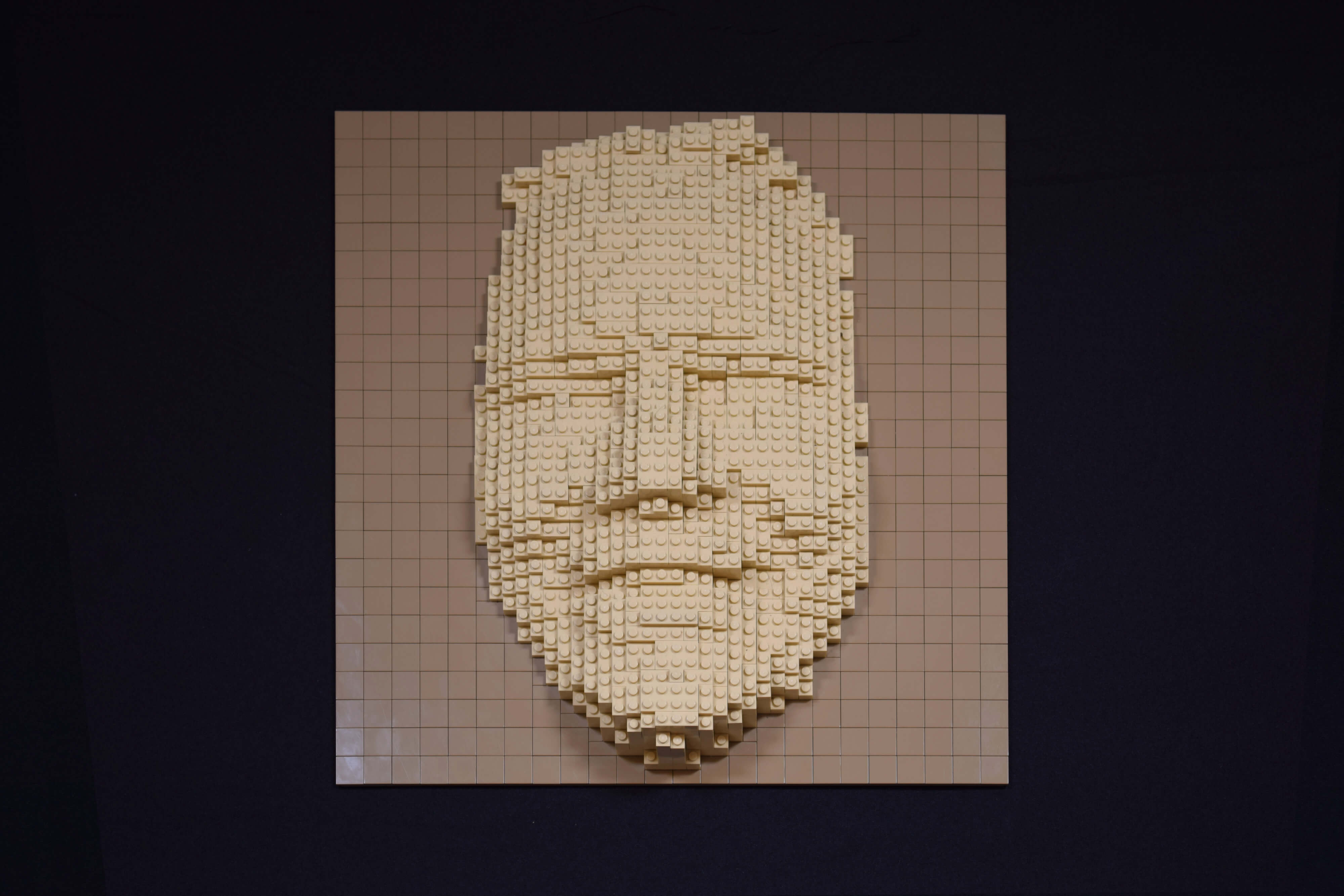Een vriendelijk lachend vrouwengezicht (of masker) gemaakt van legostenen. De sculptuur van zandsteen-kleurige stenen zijn bevestigd op een baseplate bedekt met gladde donkerbeige stenen.