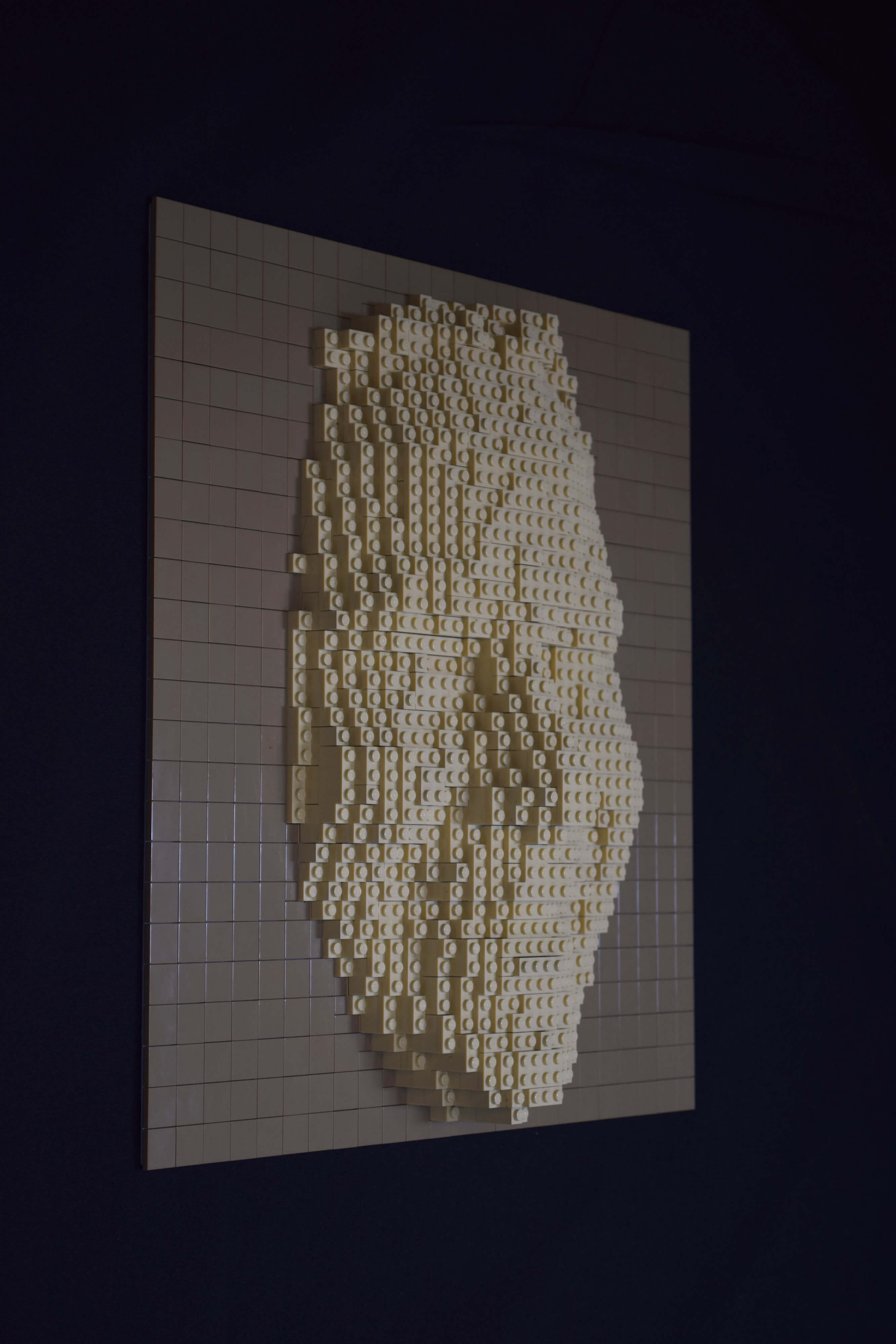 Zijaanzicht van een vriendelijk lachend vrouwengezicht (of masker) gemaakt van legostenen. De sculptuur van zandsteen-kleurige stenen zijn bevestigd op een baseplate bedekt met gladde donkerbeige stenen.