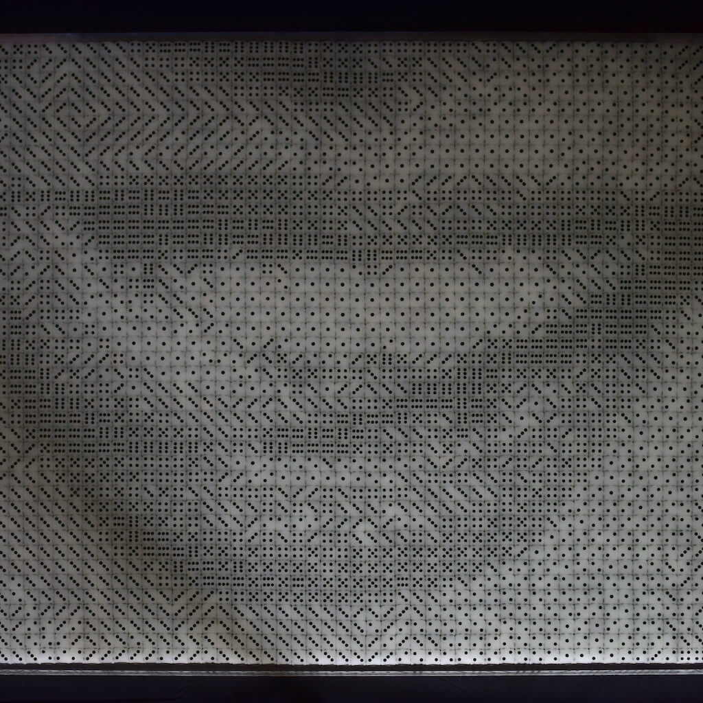 thumbnail - Dobbelstenenmozaïek van de mond van een vrouw die met het puntje van haar tong de rechterkant van haar bovenlip bevochtigd. De fosforescerende dobbelstenen hebben bij het daglicht een grijs-groene kleur en zwarte ogen.