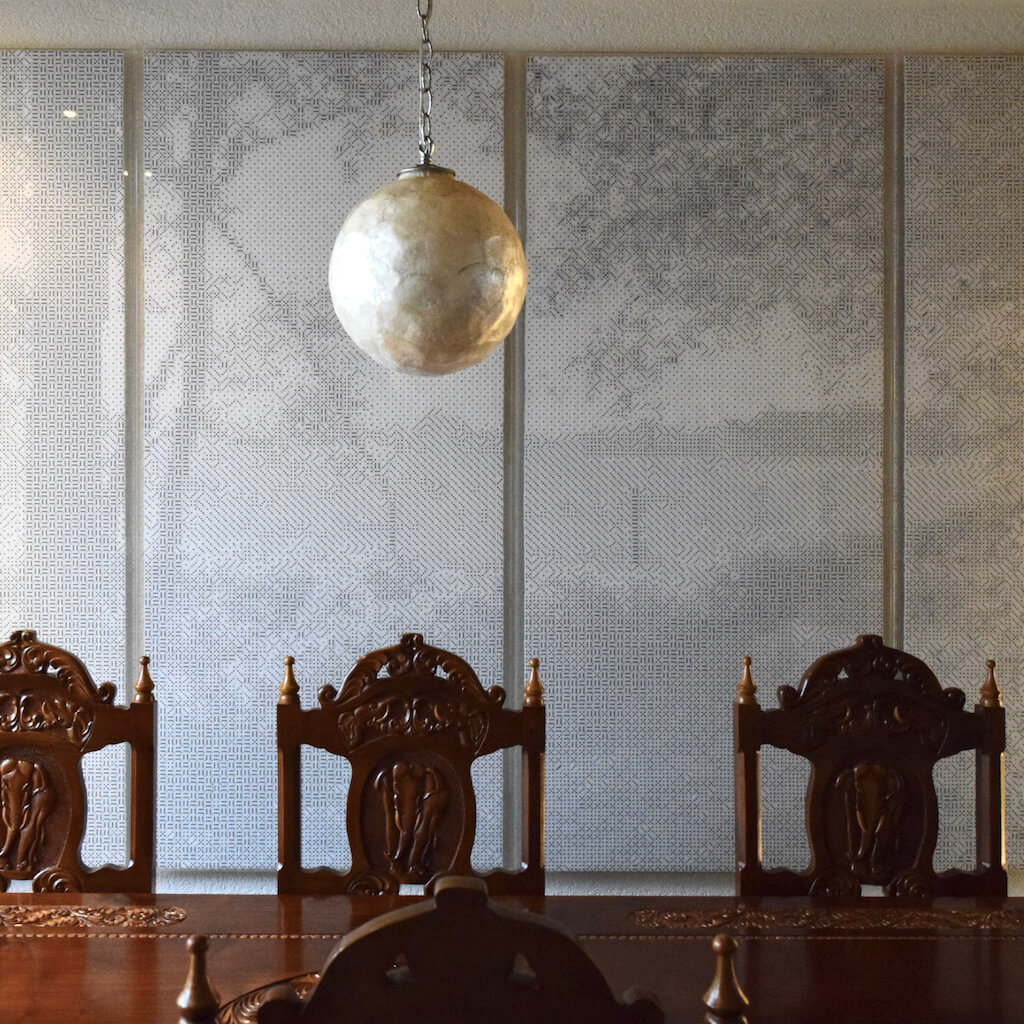 thumbnail - Een groot mozaïek van witte dobbelstenen met zwarte ogen boven een grote, klassieke, houten eettafel. Het mozaïek bestaat uit 4 langwerpige panelen, die samen een breed landschap vormen. Het onderwerp van het kunstwerk is een uitzicht over een weiland gezien vanaf een zandpad aan de bosrand. De lage zon schijnt tussen de bomen door en laat lange schaduwen van de begroeiing op het zandpad vallen.