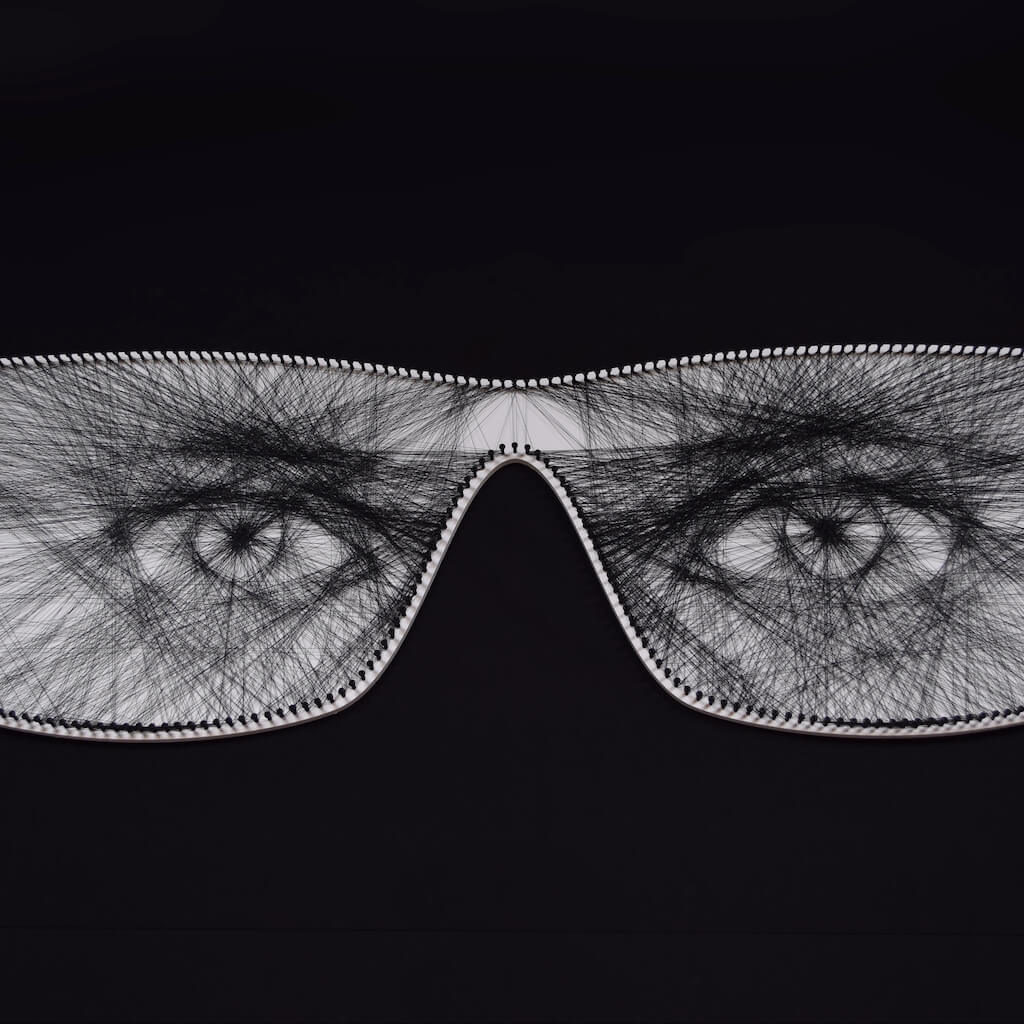 thumbnail - String art op een witte achtergrond in de vorm van een bril. De zwarte draad gespannen tussen de schroeven aan de rand van het montuur vormen de indringende blik van twee vrouwenogen.