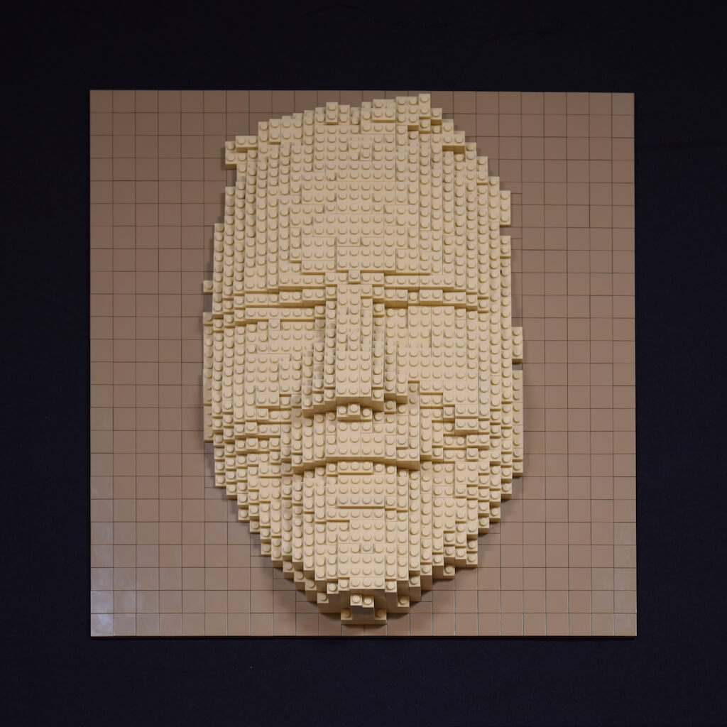 thumbnail - Een vriendelijk lachend vrouwengezicht (of masker) gemaakt van legostenen. De sculptuur van zandsteen-kleurige stenen zijn bevestigd op een baseplate bedekt met gladde donkerbeige stenen.