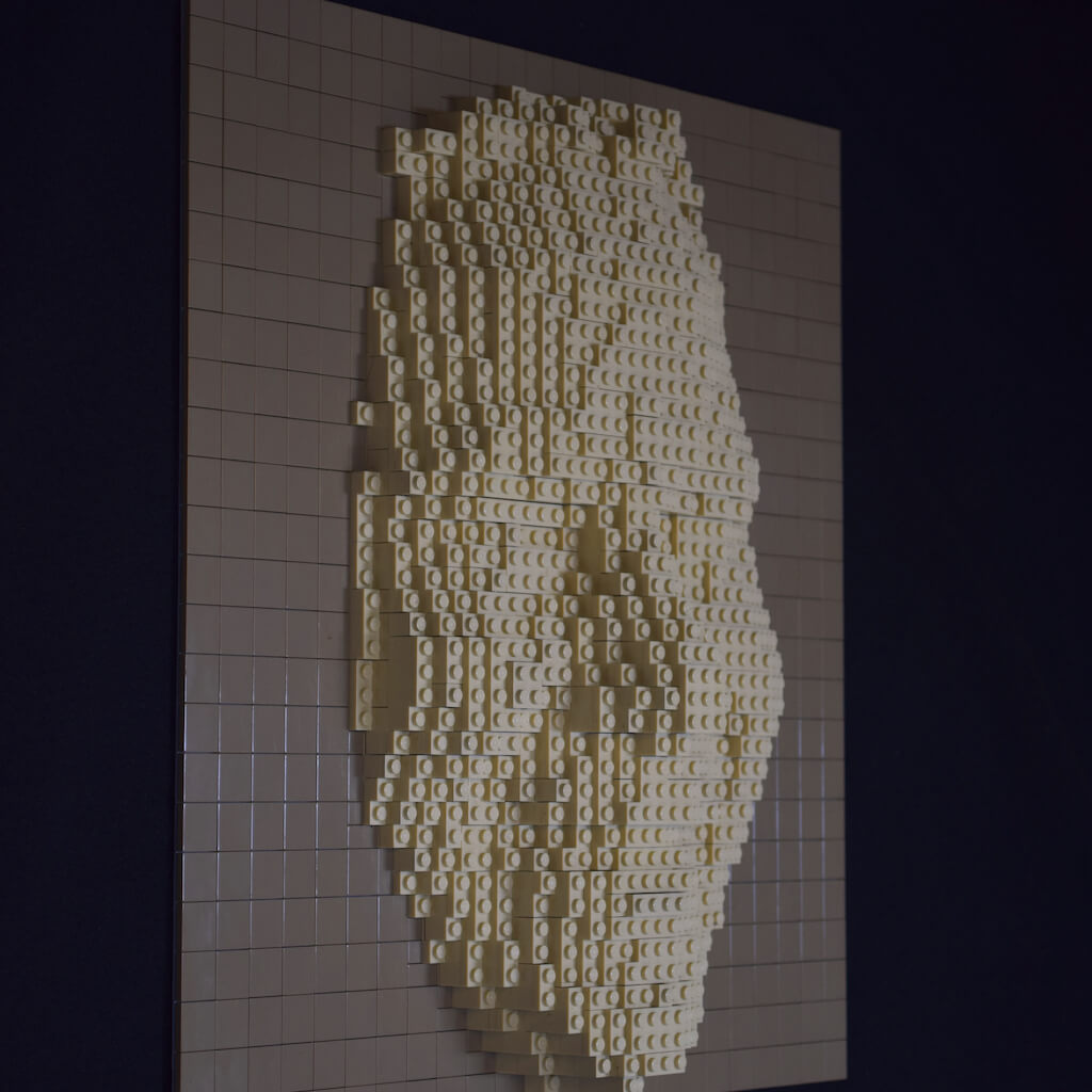 thumbnail - Zijaanzicht van een vriendelijk lachend vrouwengezicht (of masker) gemaakt van legostenen. De sculptuur van zandsteen-kleurige stenen zijn bevestigd op een baseplate bedekt met gladde donkerbeige stenen.